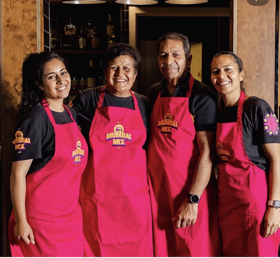 Two sisters and their parents together, wearing pink Mumbai Mix aprons